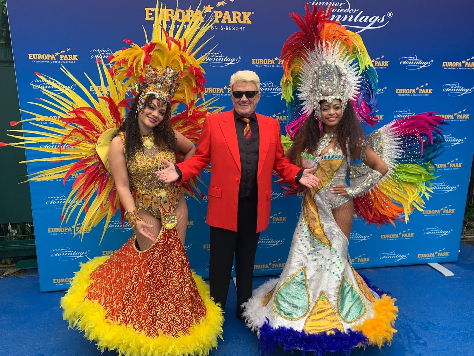 Zwei Tänzerinnen in großen Federkostümen mit einem Gast beim Fototermin vor der Europa-Park-Fotowand
